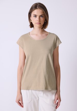 Young woman with short brown hair wearing a beige short-sleeve top with lace detail on sleeves and white pants, standing against a plain background.