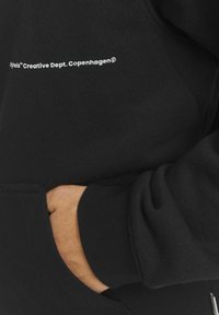 Sort hættetrøje lavet af blødt stof, med en frontlomme, ribbede manchetter og et hvidt trykt logo på siden, præsenteret mod en neutral baggrund.