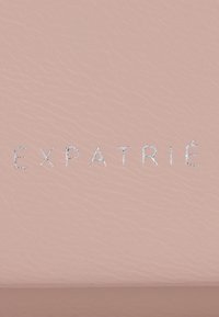 Jasnoróżowa skórka teksturowana z wytłoczonym logo "EXPATRIÉ" w kolorze srebrnym. Materiał ma subtelną poświatę i gładkie, miękkie wykończenie.