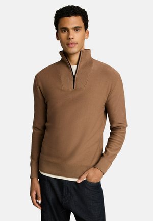 Young man wearing a brown zip-up sweater with ribbed collar and cuffs, standing with one hand in his dark jeans pocket.