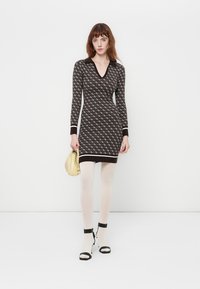 Abito in maglia con un motivo geometrico scuro su uno sfondo chiaro, scollatura a V profonda, maniche lunghe e silhouette aderente. Accentuato da una fascia nera.