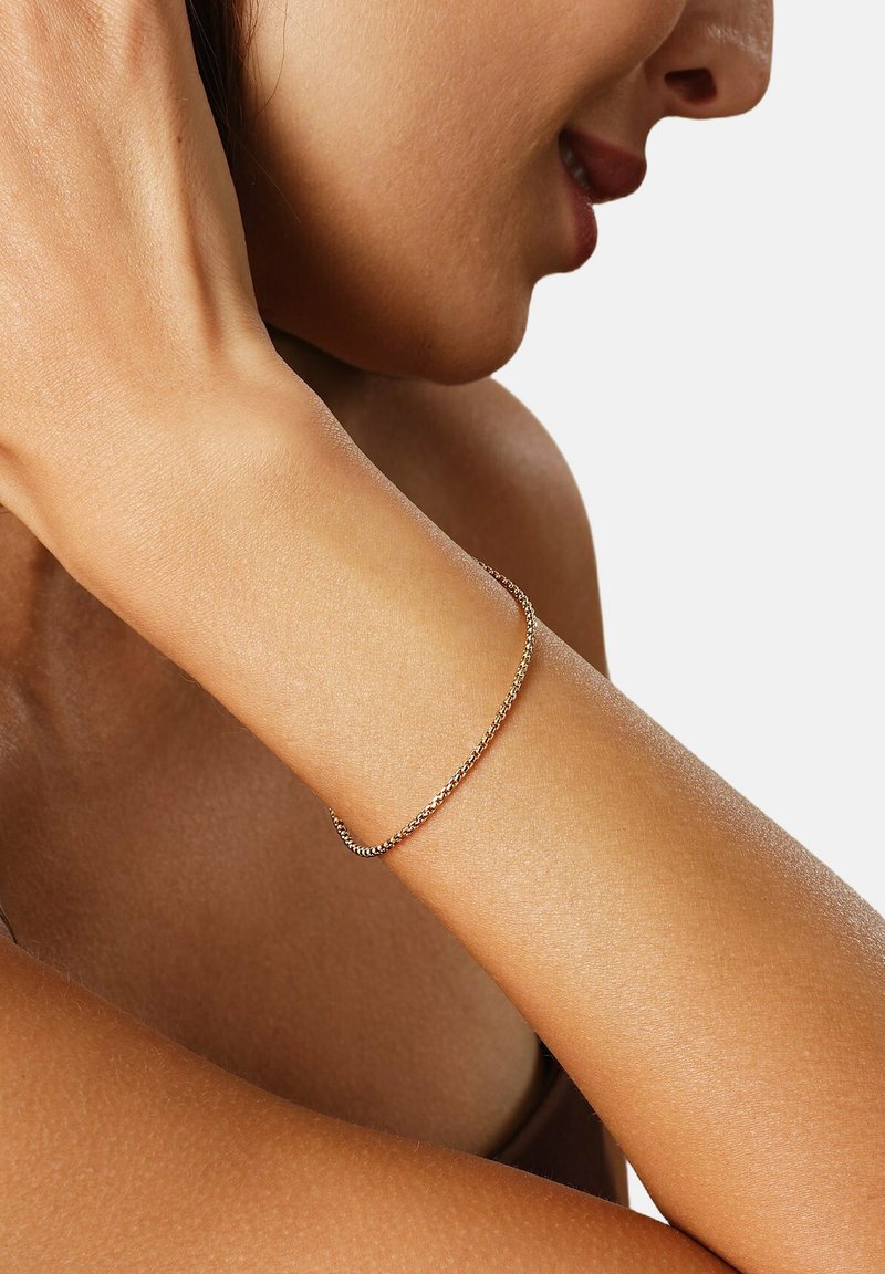 Guldtonet metalarmbånd med et delikat, snoet rebdesign, vist på et håndled mod en glat hudfarvet baggrund.