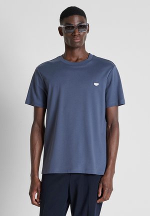 Uomo con maglietta blu scuro e occhiali da sole in piedi davanti a uno sfondo chiaro e uniforme.