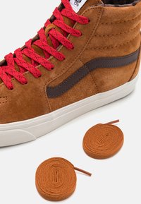 Baskets montantes en daim marron avec des lacets rouges, dotées d'une bande latérale marron foncé et d'une semelle en caoutchouc blanche. Comprend deux lacets ronds marron.