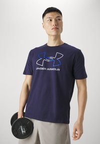 Sötétkék pamut póló rövid ujjal, amelynek elején egy feltűnő szürke és kék Under Armour logó található. A modellen kézi súlyzók vannak.