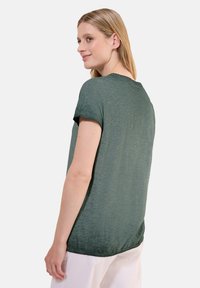 Top verde a maniche corte con una leggera lucentezza, vestibilità rilassata e scollatura rotonda. Tessuto leggero con una parte posteriore più lunga.