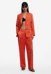 Oranje satijnen pak met een bloemenpatroon, blazer met één knoop, wijde broekspijpen en een zwarte bikini-top, aangevuld met smalle zonnebril.
