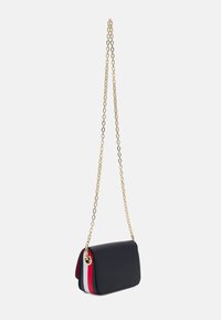 Borsa a tracolla nera con strisce laterali rosse, bianche e blu navy e tracolla in catena dorata appesa su uno sfondo bianco.