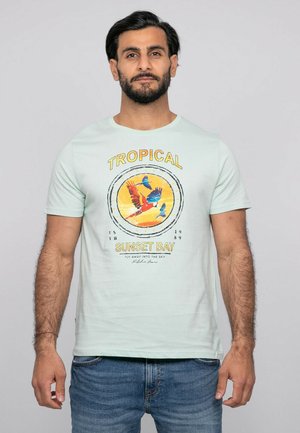 Homme portant un t-shirt bleu clair avec l'inscription "Tropical Sunset Bay" et un motif d'oiseaux colorés en vol, associé à un jean bleu, debout devant un fond gris.
