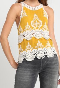 Keltainen hihaton toppi, jossa valkoiset pitsireunukset, monimutkainen kukkakuvio ja lovireunainen helma. Sileä kangas ja rento istuvuus.