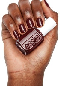 Smalto per unghie di un color bordeaux scuro in una bottiglia di vetro trasparente con un tappo bianco, tenuto in una mano con una tonalità di pelle marrone chiaro e unghie curate.