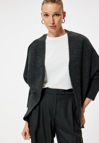 Cardigan grigio in maglia con vestibilità rilassata, apertura anteriore e maniche larghe, indossato sopra una camicia bianca e pantaloni neri su misura. Design minimalista.