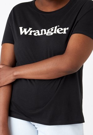 Czarna koszulka Wrangler noszona przez osobę z założonymi skrzyżowanymi ramionami, w połączeniu z jasnymi spodniami, na jednolitym tle.