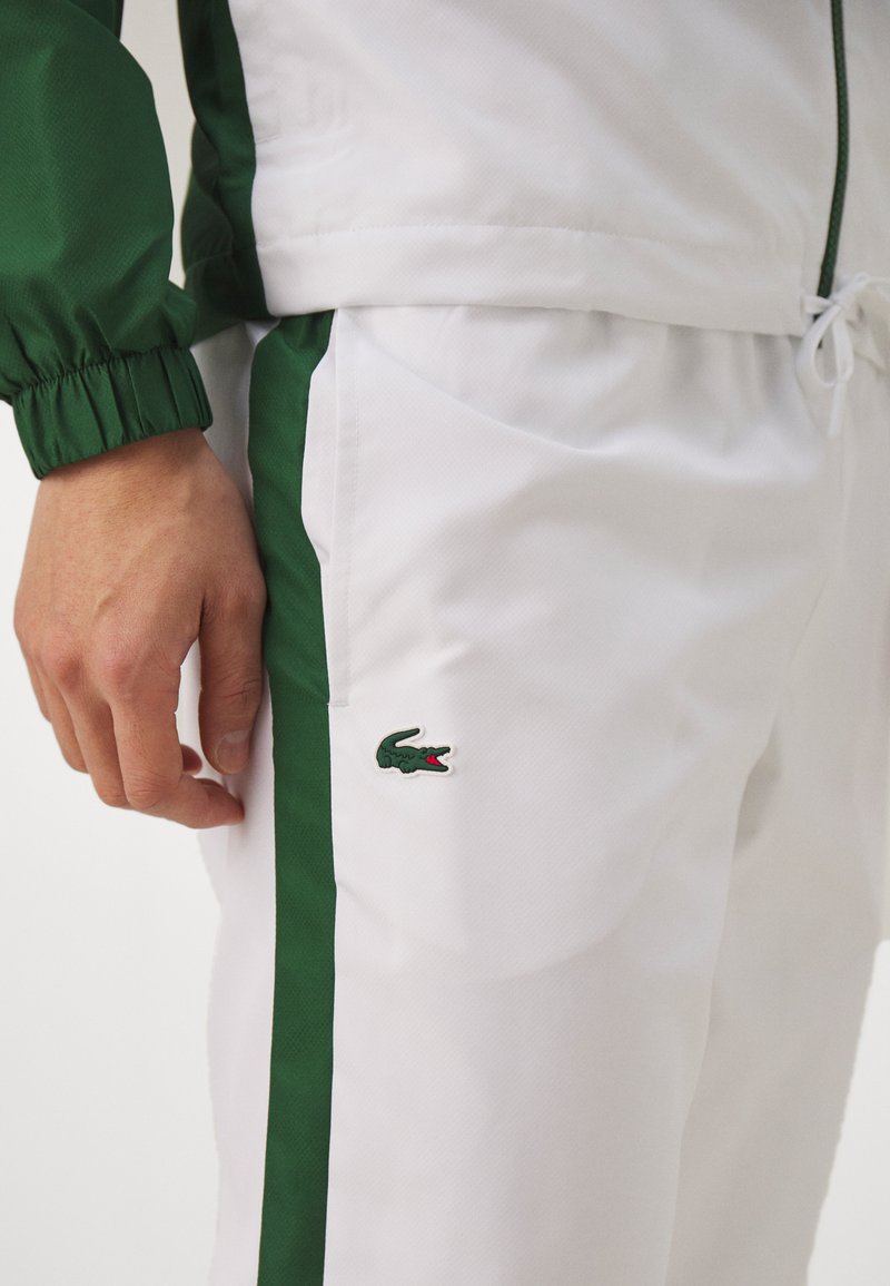 Pantalones de chándal blancos con rayas laterales verdes, cinturilla elástica y el logo de Lacoste destacado en el muslo. Tejido ligero y suave.