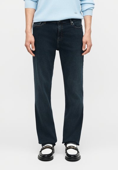 Άνδρας φορώντας σκούρο μπλε τζιν ίσιας γραμμής, λευκές κάλτσες, μαύρα και λευκά μοκασίνια και ανοιχτό μπλε ριπ πουλόβερ.