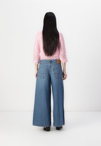 Kvinna med långt mörkt hår som bär en rosa stickad topp och vida blå denimjeans, stående mot en vit bakgrund, vänd bort.