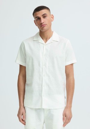 Jeune homme aux cheveux courts portant une chemise blanche à manches courtes et un pantalon blanc, sur un fond clair uni.