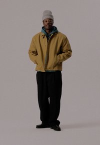 Κίτρινο βαμβακερό μπουφάν πάνω από μια τιρκουάζ μπλούζα, συνδυασμένο με μαύρες φαρδιές παντελόνες και γκρίζο πλεκτό σκουφάκι. Μαύρα παπούτσια ολοκληρώνουν την εμφάνιση.