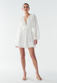 Vit klänning med långa ärmar, djup v-ringning och insvängd midja. Genombruten texturerad tyg med subtilt mönster. Buren med beige sandaler med klackblock.