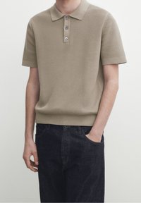 Beige texturoitu lyhythihainen poolopaita, jossa on kolmipainikkeinen nappilista ja resorinen helma, yhdistetty tummiin farkkuihin.