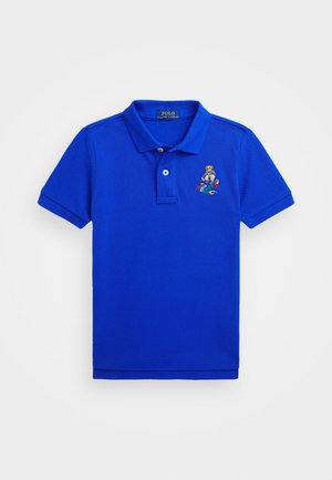 Koszulka polo w kolorze królewskim niebieskim z krótkim rękawem, dwoma guzikami, kołnierzykiem i małym wyszywanym misiem trzymającym deskorolki na lewej piersi.