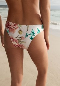 Spodnji del bikinija z rožnim vzorcem ima belo osnovo z velikimi rožnatimi in zelenimi cvetovi. Material je videti gladek, s preprostimi linijami in minimalnim šivanjem.