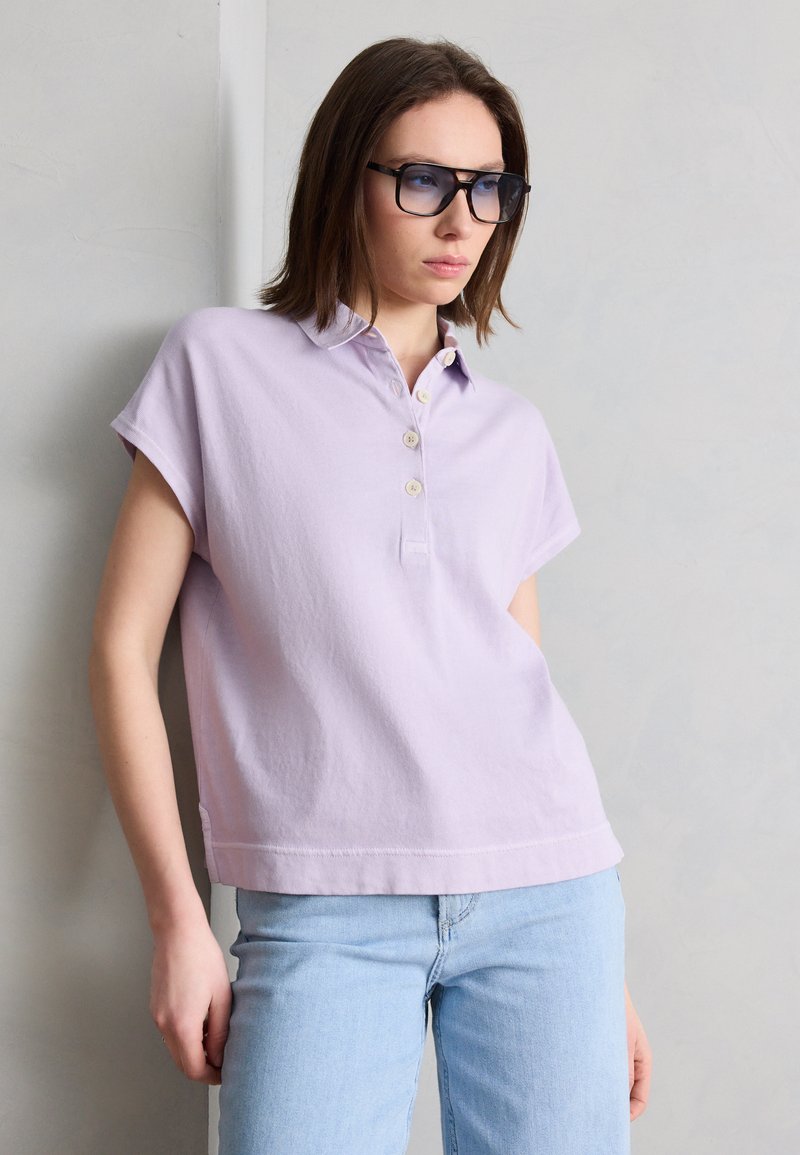 Νεαρή γυναίκα που φοράει μαύρα γυαλιά ηλίου, ένα ανοιχτό μοβ πουκάμισο με κοντά μανίκια και κουμπιά και ανοιχτό μπλε τζιν, στηριζόμενη σε έναν άσπρο τοίχο.