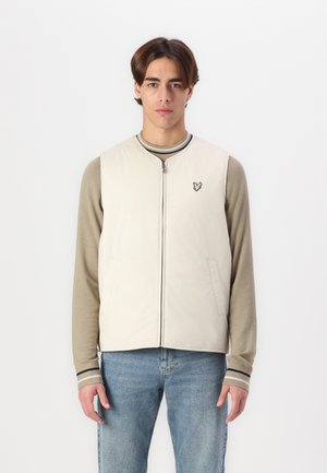 Vest in light beige, zip-up design with two side pockets, featuring a small embroidered logo on the chest and paired with a long-sleeve top.
