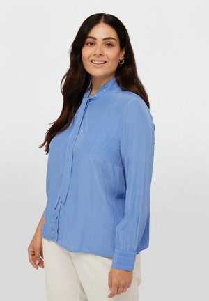 Blusa azzurra chiara realizzata in tessuto morbido, con colletto decorato da piccoli chiodini, maniche lunghe e chiusura con bottoni davanti.