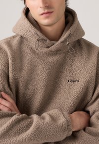 Brown fleece hoodie with a textured finish, drawstring hood, and black "Levi's" logo on the chest. Soft and loose-fitting design.