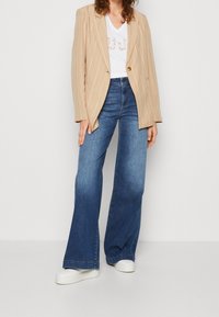 Kvinna i beige randig kavaj, vit t-shirt med grafik, högmidjade vida blå jeans och vita plattformssneakers som står mot en enfärgad vägg.