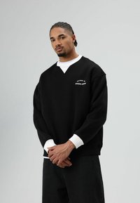 Zwarte trui met V-hals en witte manchetten, met "STUDIO ARTS" in witte letters op de borst. Het model draagt donkere spijkerbroek.