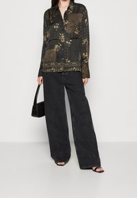 Blusa nera a maniche lunghe con motivo e dettagli floreali, dotata di due tasche sul petto, abbinata a jeans neri a gamba larga e sandali con cinturini.