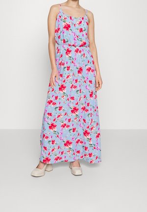 Vrouw die een mouwloze lichtblauwe maxi-jurk draagt met een rood- en roze bloemenpatroon en beige pumps met blokhak.