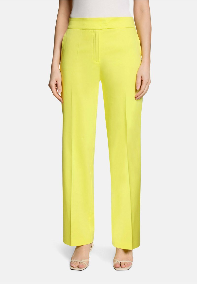Femme portant un pantalon large jaune vif et des talons blancs à brides, debout, les mains détendues le long du corps.