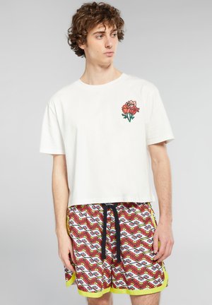 T-shirt cropped bianco con una grafica di una rosa sul petto, abbinato a pantaloni corti colorati con motivo geometrico e un laccetto nero.