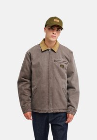Jeune homme portant une veste marron zippée avec un col beige et une casquette de baseball vert olive, debout devant un fond uni clair.