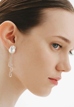 Mujer de piel clara y cabello oscuro lleva un gran pendiente de perla unido a tres gotas de cristal colgantes sobre un fondo blanco.