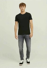 Young man standing, wearing a plain black t-shirt, faded gray jeans, and black and white sneakers on a neutral background.