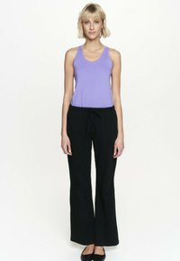 Blondynka stojąca przodem, ubrana w fioletowy top bez rękawów, czarne szerokie spodnie i czarne płaskie buty.