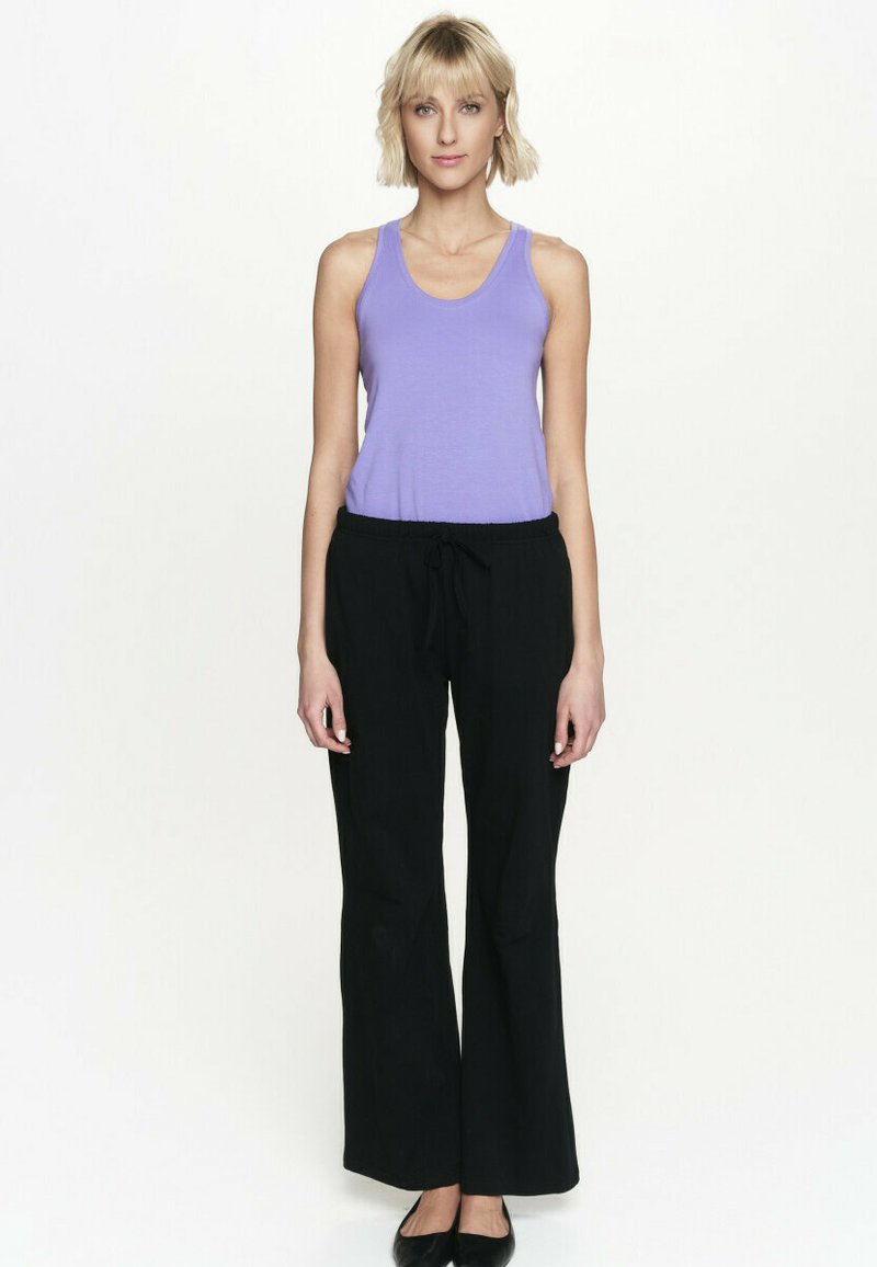 Blondynka stojąca przodem, ubrana w fioletowy top bez rękawów, czarne szerokie spodnie i czarne płaskie buty.