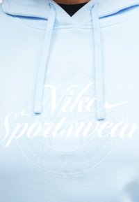 Svetlomodrá mikina s kapucňou s razeným obručovým logom, srdcovými šnúrkami a výrazným bielym textom "Nike Sportswear" na prednej strane.