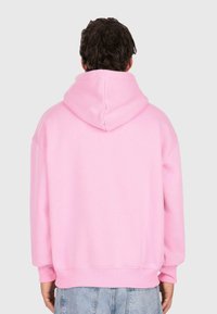 Mann mit dunklem, lockigem Haar, der einen schlichten rosa Hoodie und blaue Jeans trägt, stehend mit dem Rücken zu einem schlichten grauen Hintergrund.