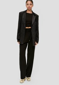 Zwarte glinsterende blazer over een zwarte cropped top, gecombineerd met losse zwarte broek en glanzende gouden hakken. Het gelikte haar maakt de look af.