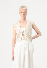 Beige knitted vest with diamond pattern, short sleeves, and front tie detail. Worn over a white top and white high-waisted skirt.