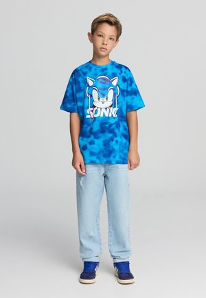 T-shirt tie-dye blu con grafica di Sonic the Hedgehog, abbinata a jeans di denim chiaro e sneaker navy. Stile casual e vestibilità comoda.