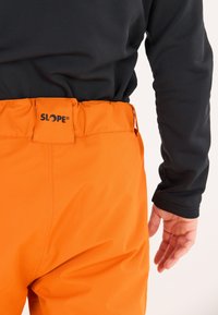 Orange Skihose mit Taschen, elastischem Bund und schwarzem Logoetikett. Schwarzes Langarmshirt teilweise im Hintergrund sichtbar.