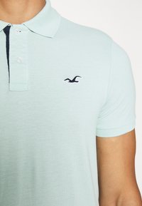 Gros plan d'une personne portant un polo à manches courtes bleu clair avec un petit logo d'oiseau sombre sur la poitrine.