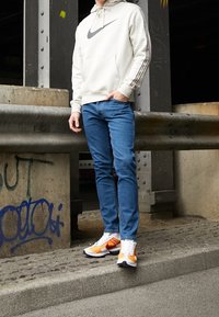 Person som står på en kantbrygga iförd en vit Nike-hoodie, blå jeans och orangea samt vita sneakers nära en betongsperre prydd med graffiti.