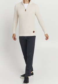 Kermanvärinen, teksturoitu neulepaita, jossa on puolivetoketjullinen kaulus, resorit hihansuissa ja ruskea logoplässi, yhdistetty tummiin housuihin ja mustiin tennareihin.